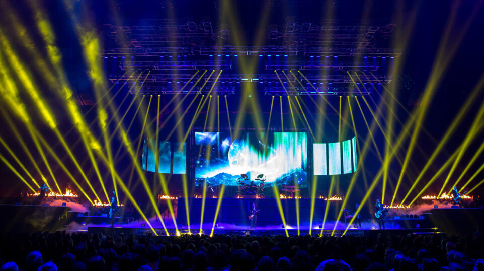 a blue stage lit up with yellow lights as a band performs to a packed audience