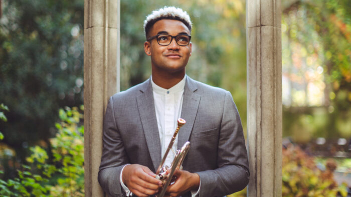 Aaron Azunda Akugbo holds a trumpet against a stone archway