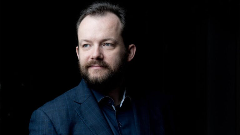 Conductor Andris Nelsons smiles in a dark suit in front of a black background