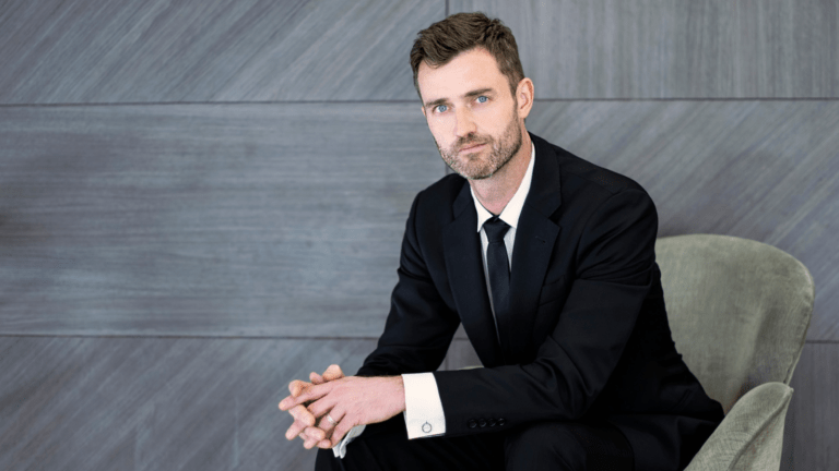 Portrait of Aaron Sheehan in dark suit, white shirt, dark tie, seated.