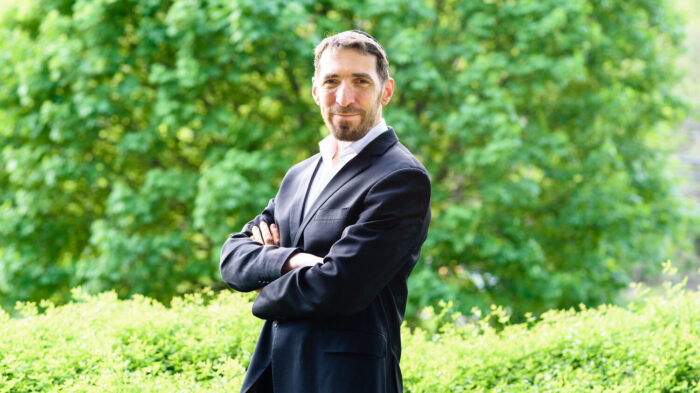 Portrait of Jonathan Leshnoff, dressed in a suit, in front of bushes and a tree