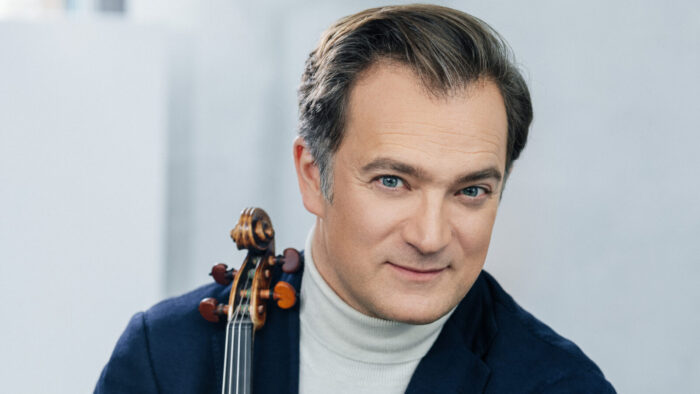 Renaud Capuçon, holding a violin and sporting a white turtleneck and blue sportcoat, gives a half smile