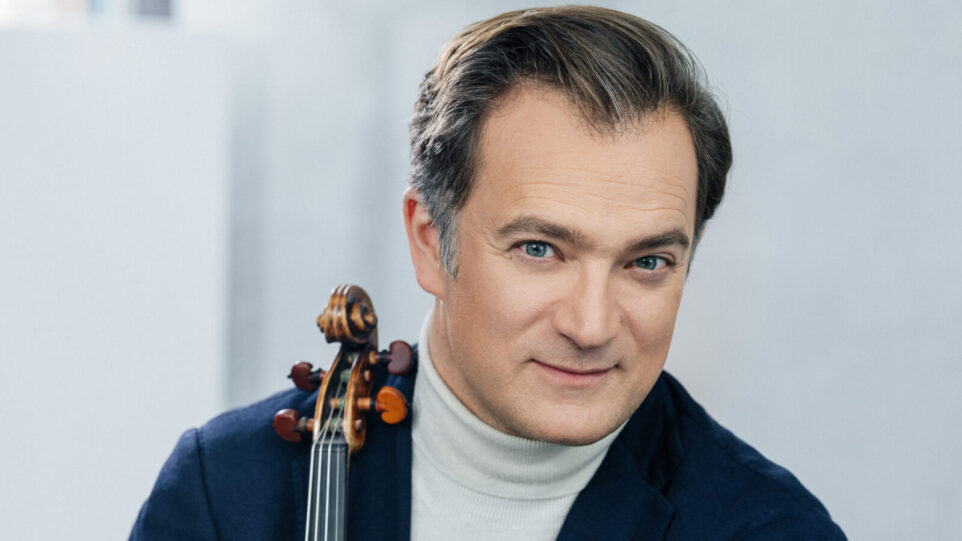 Renaud Capuçon, holding a violin and sporting a white turtleneck and blue sportcoat, gives a half smile