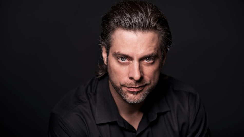 Portrait of John Relyea, brooding, staring directly into camera, dark hair slicked back, in black shirt against dark background.