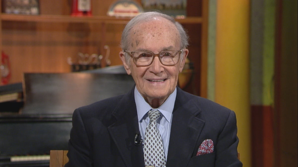 Newt Minow smiles at the camera on the set of WTTW's Chicago Tonight wearing a suit, tie, and glasses