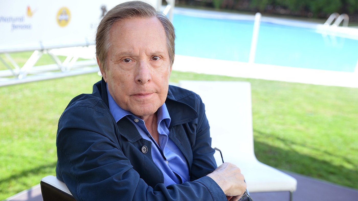 William Friedkin, dressed in a blue oxford and coat, sits outside, looking over his shoulder at the camera