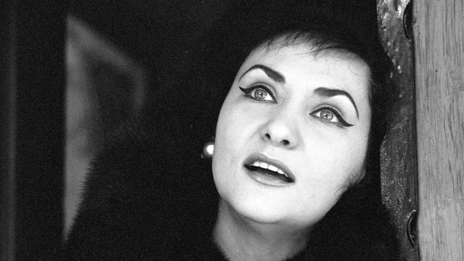 Virginia Zeanni, dressed glamorously in a fur and earrings, poses against a wall