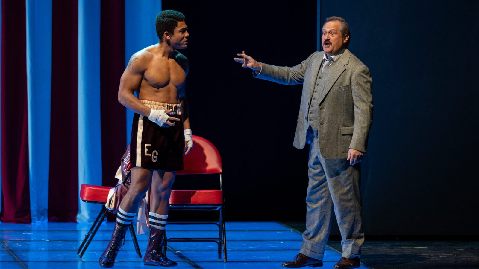 Justin Austin, shortless and in boxing shorts, performs alongsided Paul Groves, in a suit, who gesticulates towards Austin while holding a cigar