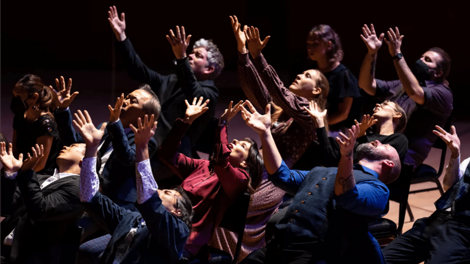 12 singers in business attire leaning back on black chairs, their arms outstretched to the sky; tormented facial expressions.