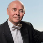 Jorge Federico Osorio gives a soft smile at a piano in front a big blue sky.