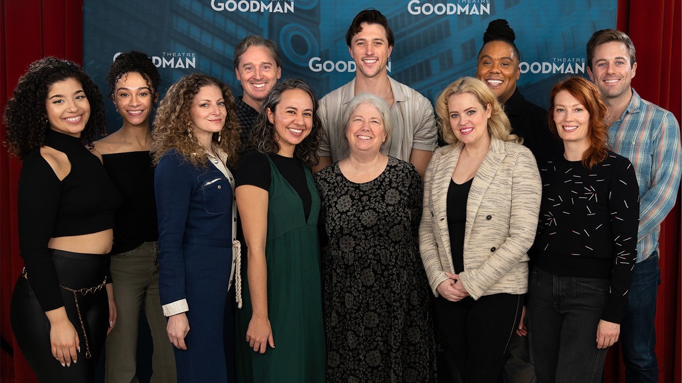 Eleven artists — including Mary Zimmerman — pose together in front of a red curtain and a Goodman Theatre sign in between rehearsals