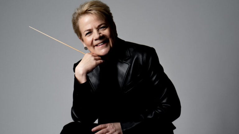 Marin Alsop sits cheerfully, wearing a black leather jacket and holding a conductors baton against a white backdrop