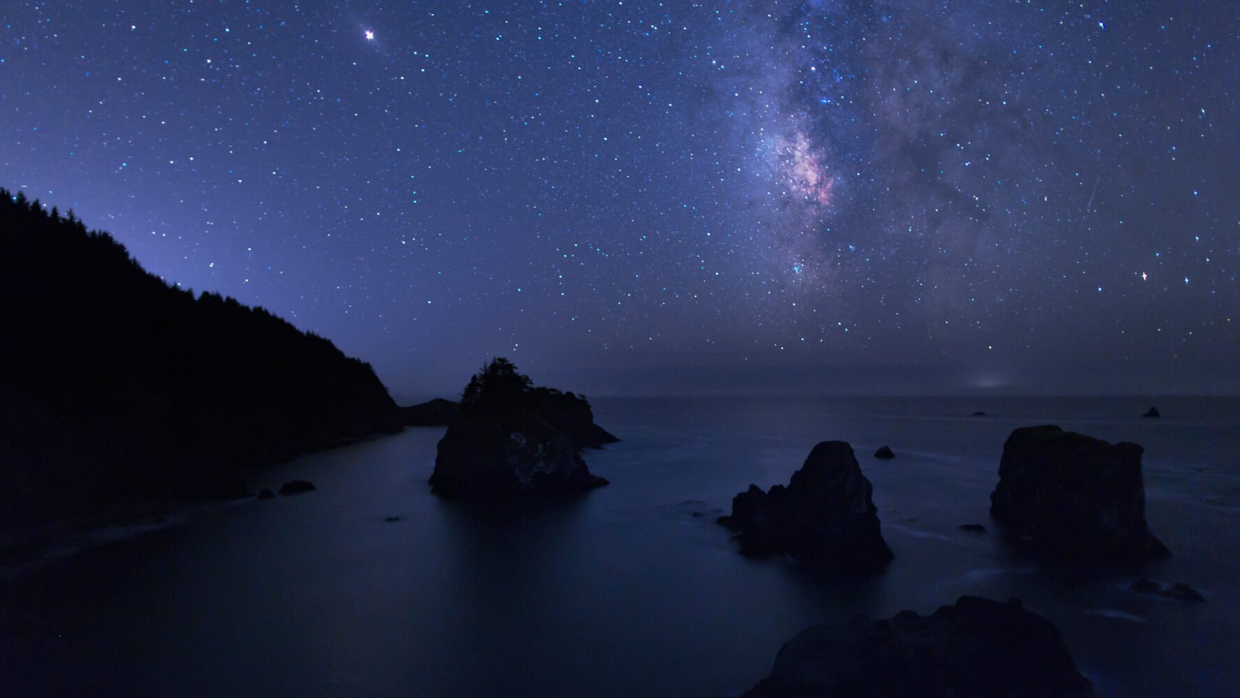 outcrops of cliffs and boulders line a sandy beach, all illuminated by the starry night sky