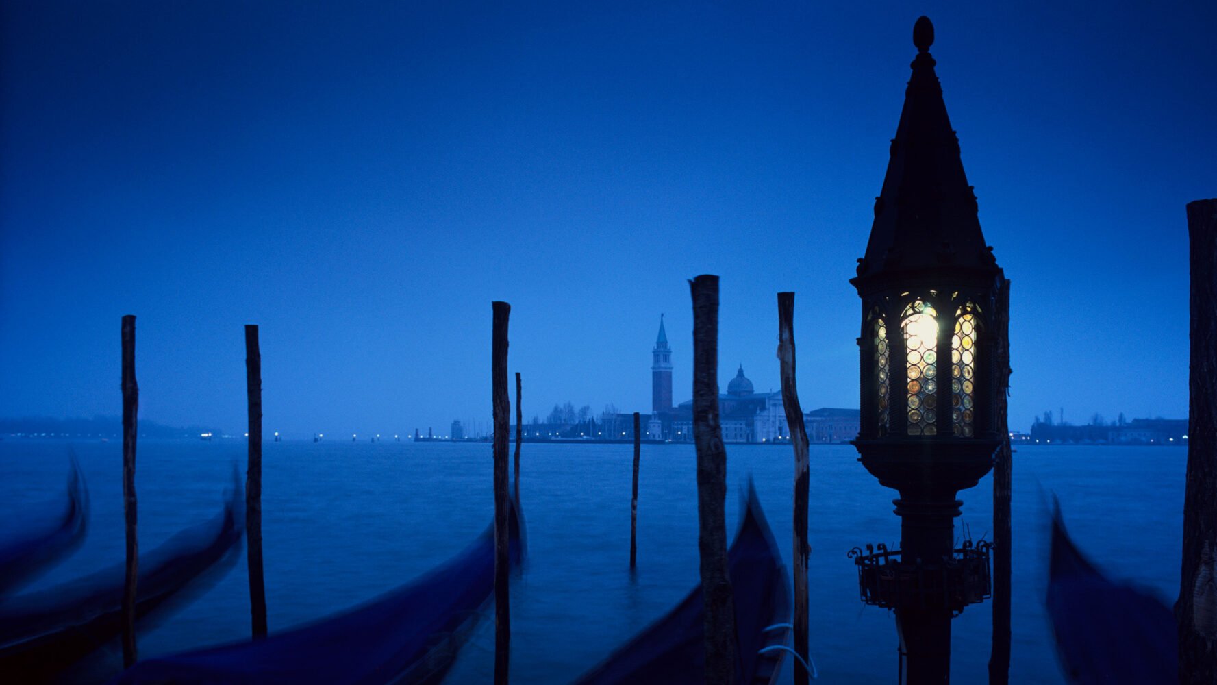 a lit lantern and vacant gondolas sit at night on a Venice canal with the Saint Mark's Bell Tower in the distance