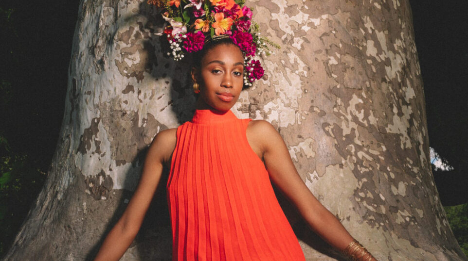Jazzmeia Horn, wearing a flowery headdress and a bright orange-red dress, stands in front of a tree trunk