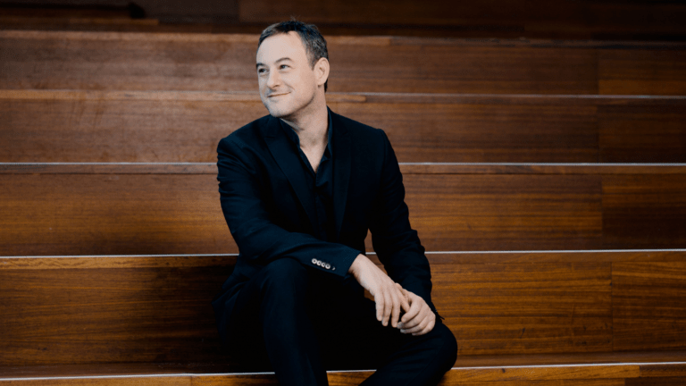 Jonathan Cohen in black suit, black shirt, open collar, sitting on wood steps that ascend past frame of image, looking over his right shoulder smiling.