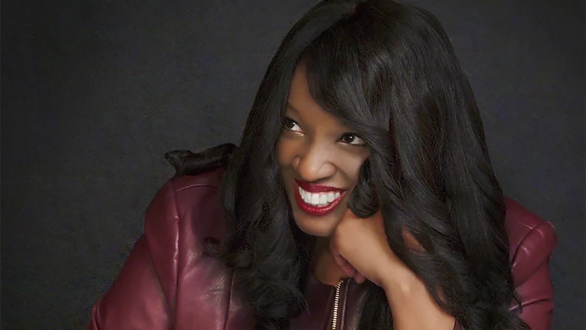 Nkeiru Okoye smiles, wearing a plum leather jacket, against a grey background