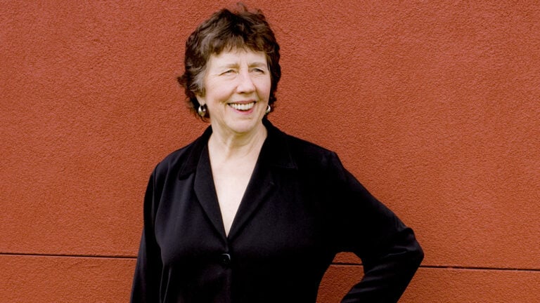 Joan Tower smiles, dressed in black, posing against a brick red stucco wall