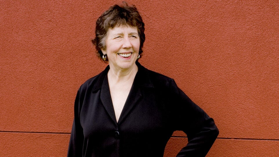 Joan Tower smiles, dressed in black, posing against a brick red stucco wall