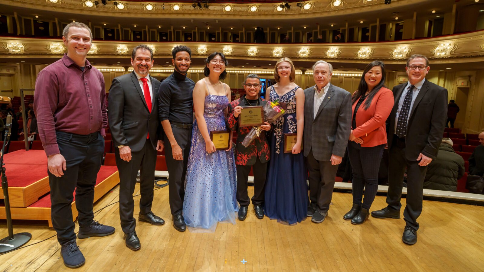 A group of 9 people pose onstage at Symphony Center, including Robbie Ellis, prizewinner Jaden Teague-Núñez, horn player Elyse Schlesinger, and flute player Chloe Nam