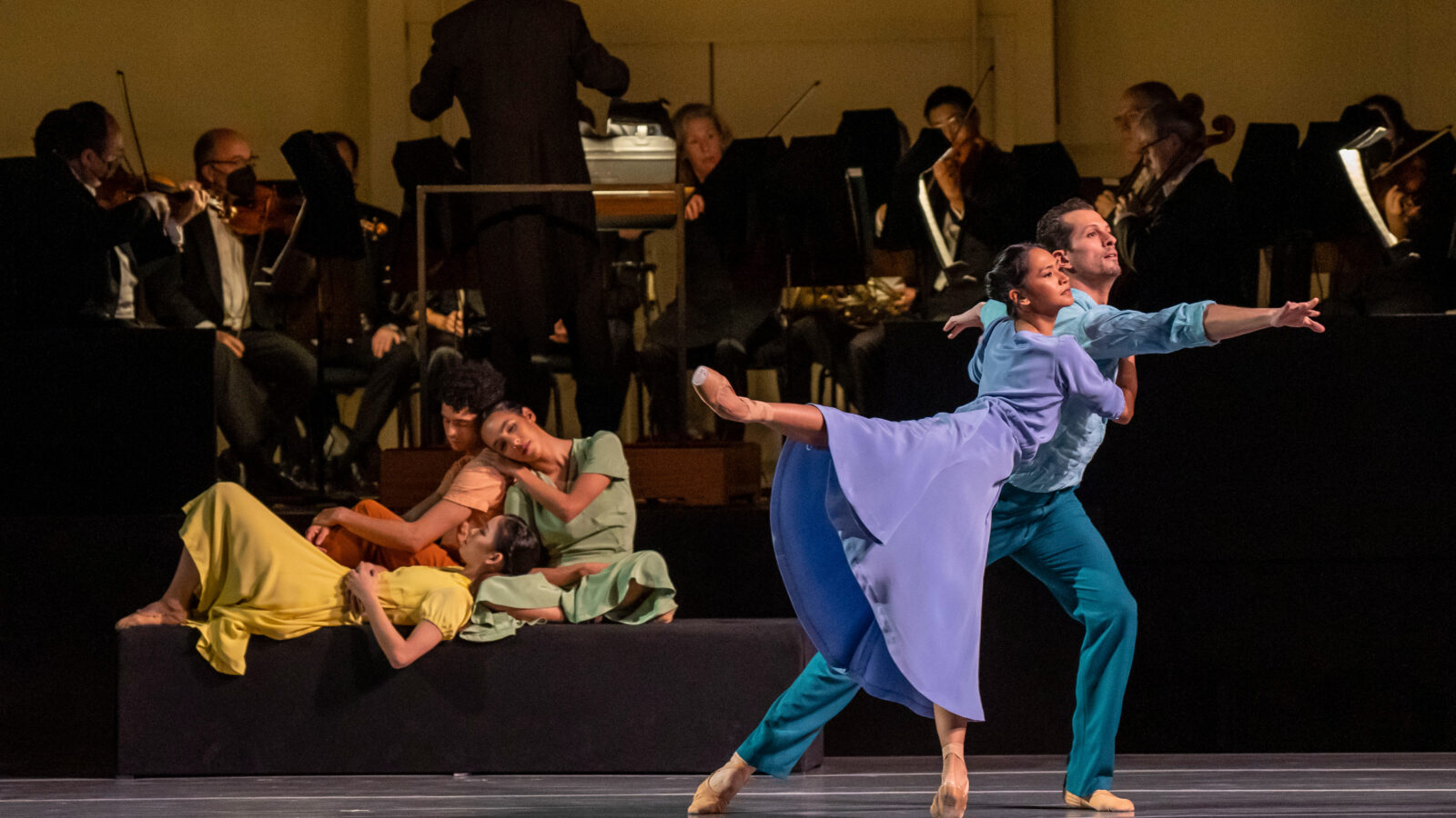 brightly dressed dancers move onstage with orchestra members in the background