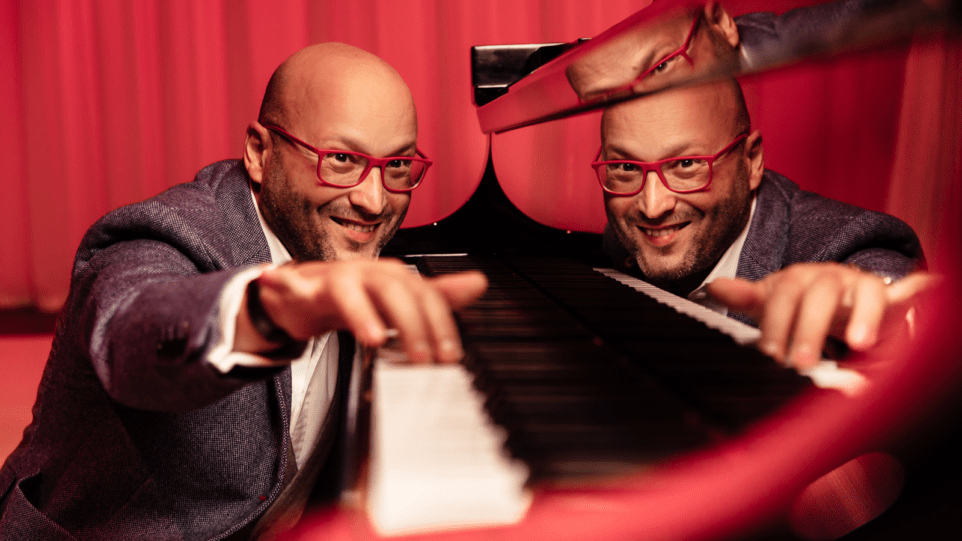 Whimsical image of Enrique Mazzola in a grey sportscoat, white shirt, no tie, red-framed eyeglasses, at the piano, crouching so that keyboard is at eye level, his reflection visible on the shiny black surface of the piano, red curtain as background.