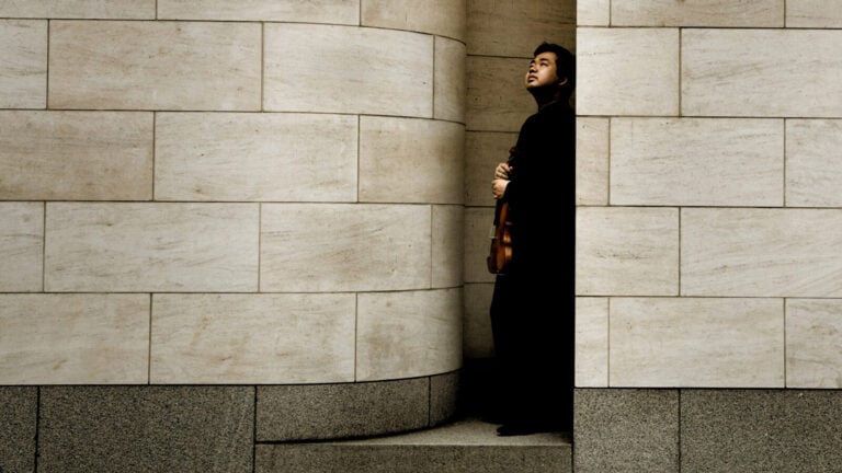 Ning Feng leans against a stone wall, holding a violin, gazing pensively to the sky