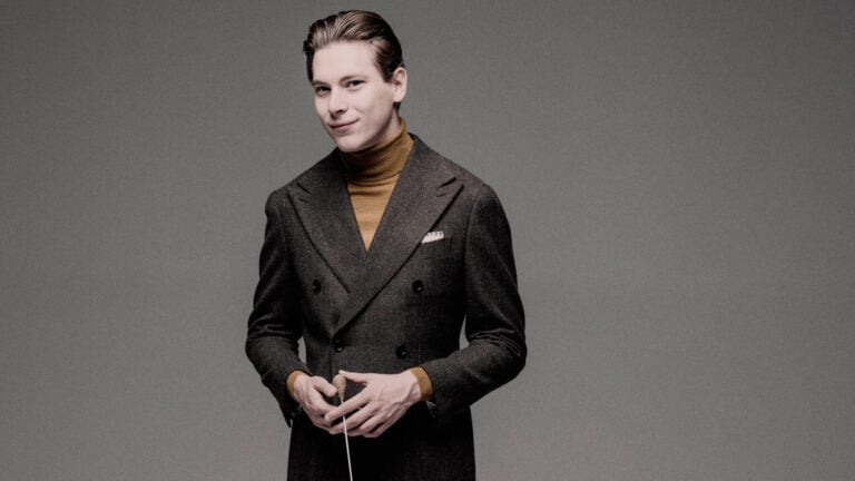 Klaus Mäkelä, dressed in a wool suit and brown turtleneck, gives a half smile while holding a conductor's baton