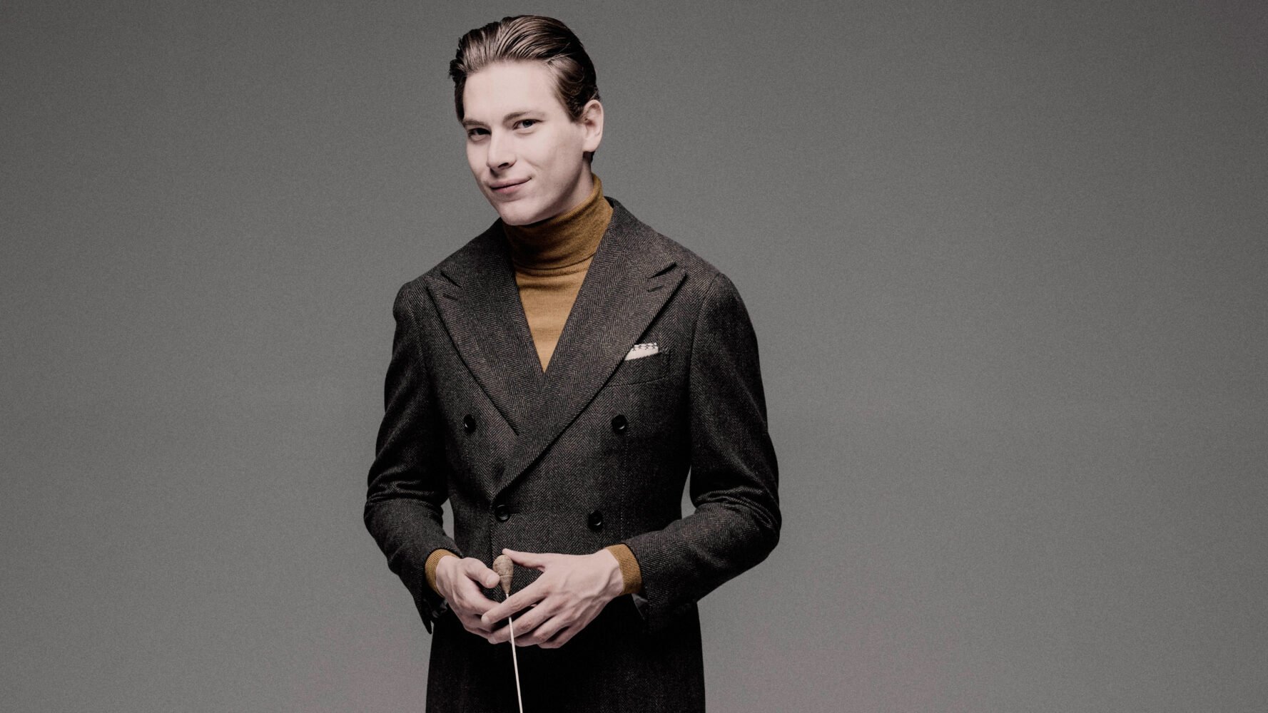 Klaus Mäkelä, dressed in a wool suit and brown turtleneck, gives a half smile while holding a conductor's baton