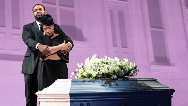 A tall African-American man hugs a shorter African-American woman from behind. The woman is crying. The man is looking off into the distance. They are standing by a white, closed casket with flowers on top.