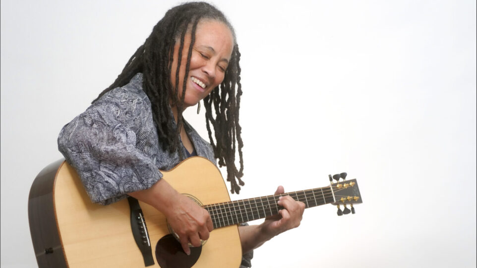 Black woman with long braids smiles and plays guitar