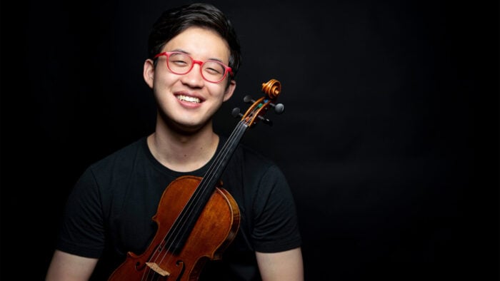 Julian Rhee smiles wearing red eyeglasses and black tee shirt swhile holding a violin