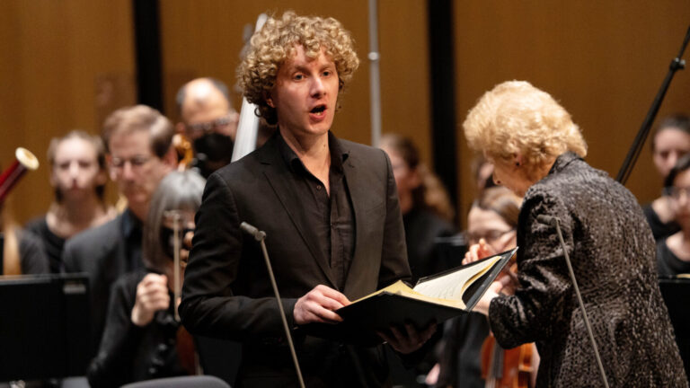 Gwilym Bowen singing onstage in black suit, with conductor and musicians in background