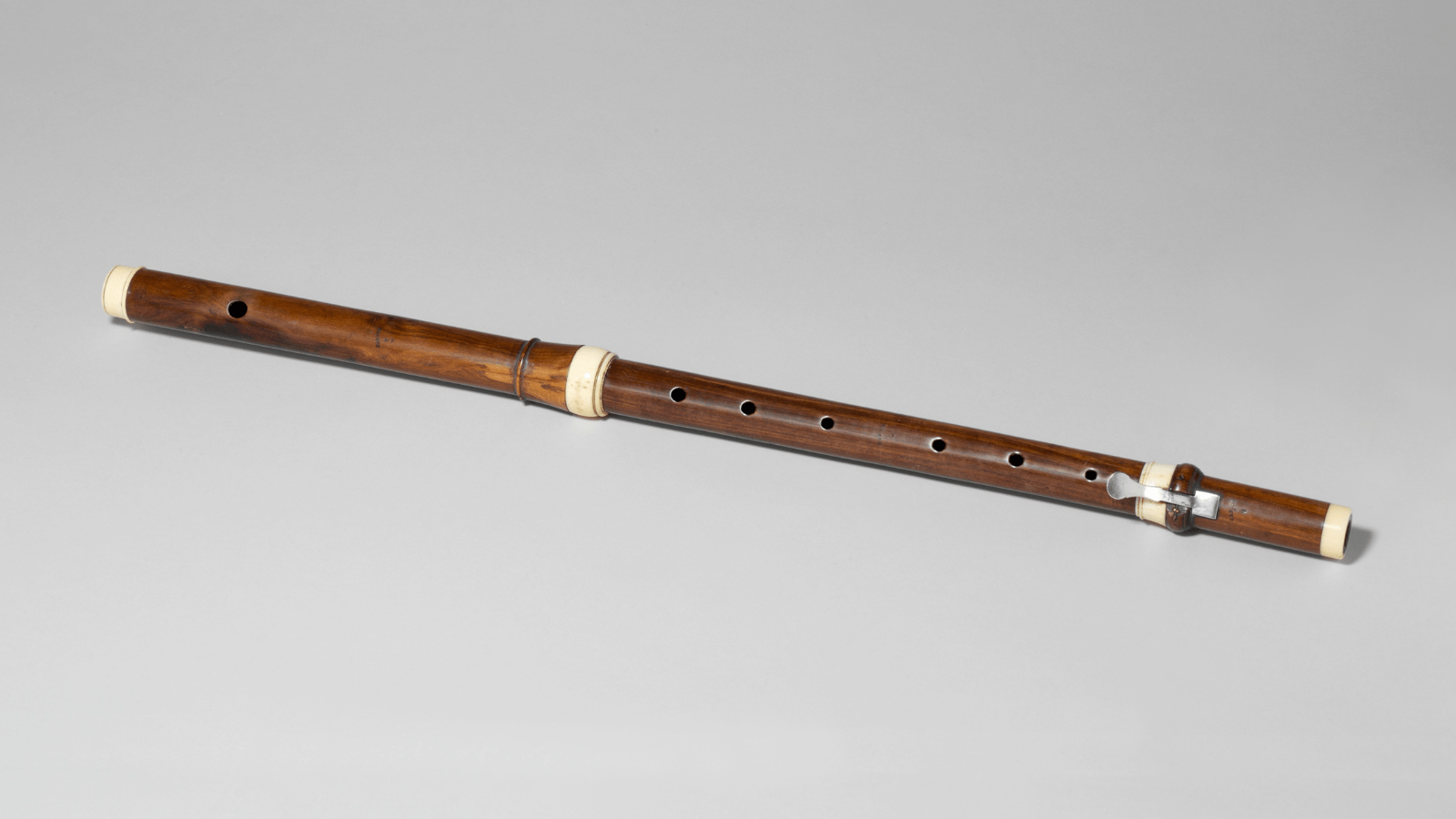 Picture of a wooden, transverse flute, estimated to be from the years 1720-1740. The flute is against a plain, light grey background. 
