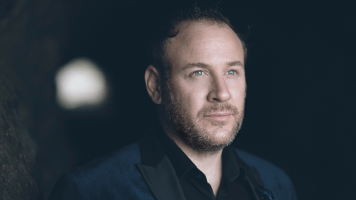 Lucas Meachem in navy tuxedo with dark shirt, open collar, soft lighting, dark background.