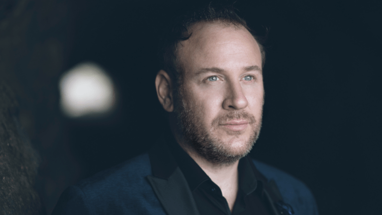 Lucas Meachem in navy tuxedo with dark shirt, open collar, soft lighting, dark background.