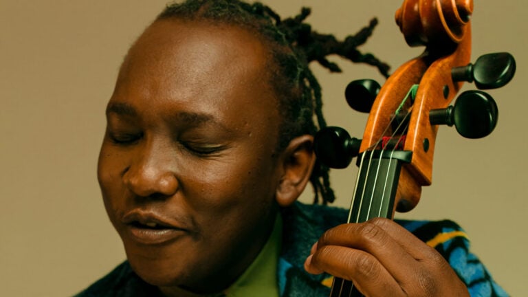 Abel Selaocoe, in a vibrant blue suit and green shirt, is immersed in music as he plays the cello with eyes closed