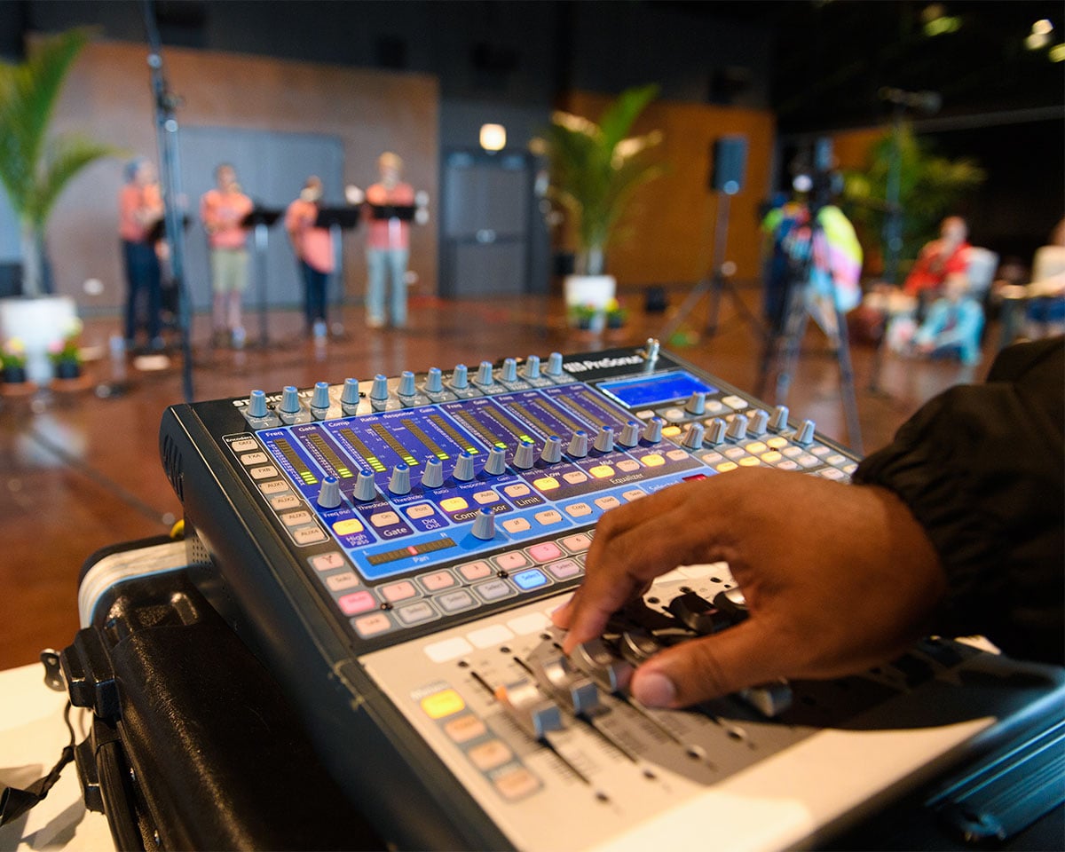 Hand operating knobs on sound board with performers in background, blurred