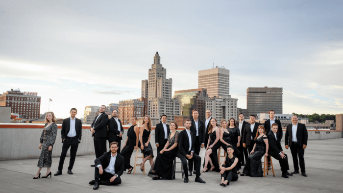 22 members of choir; men in black suits, white shirt, no tie; women in black dresses; posing on a rooftop in Providence, Rhode Island with the city's skyline as the background.