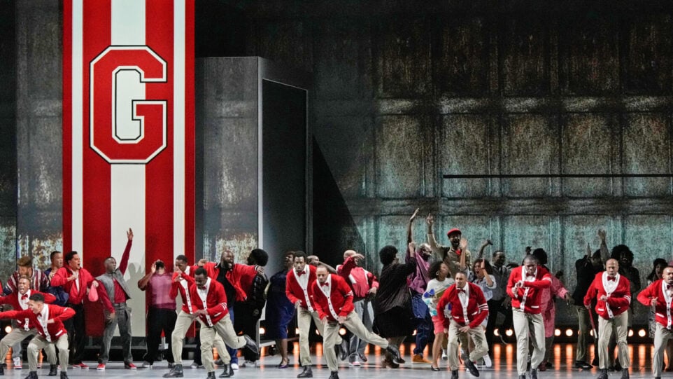 A group of dancers in motion wear letterman bright red letterman cardigans in front of a red banner with the letter "G"