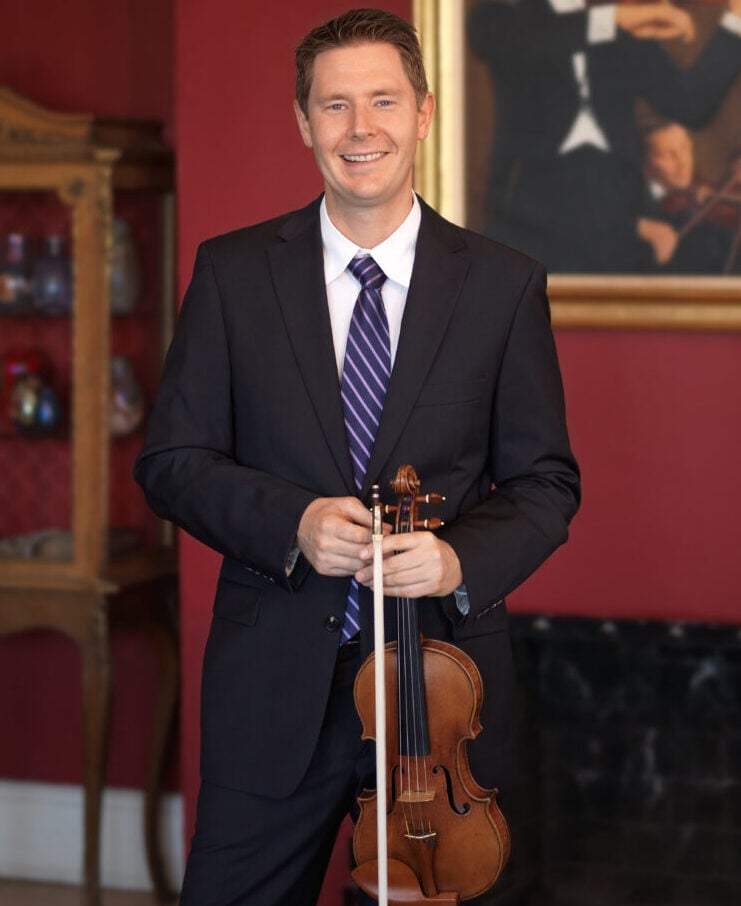 A picture of John Gerson, Senior Sales Executive at Bein & Fushi. He is a white man in his forties with brown hair and blue eyes. He is smiling at the camera while hold a violin loosely by the scroll.