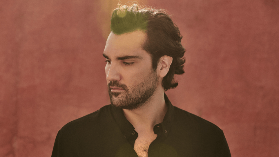 Overexposed portrait of Jonathan Tetelman in profile wearing an open-collared dark shirt in front of a red stone wall.