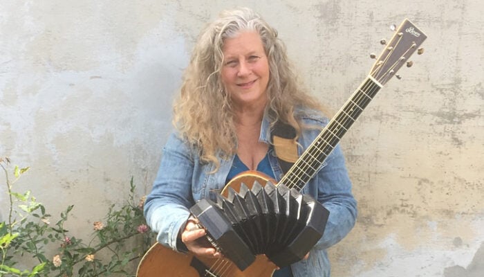 smiling woman with long gray hair holds a guitar and a concertina