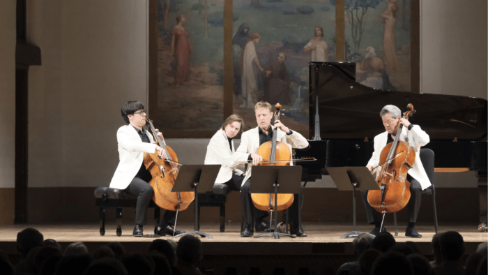 A group of four men, all in white, are playing a piece of chamber music. The group consists of a 3 cellists and a pianist.