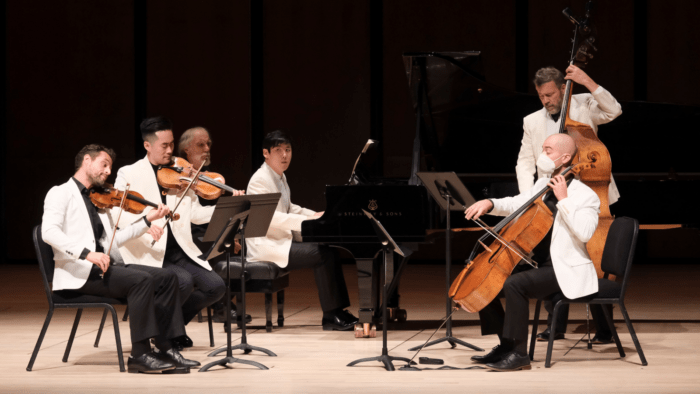 A group of five men, all in white, are playing a piece of chamber music. The group consists of a violinist, violist, cellist, double bassist, and a pianist.