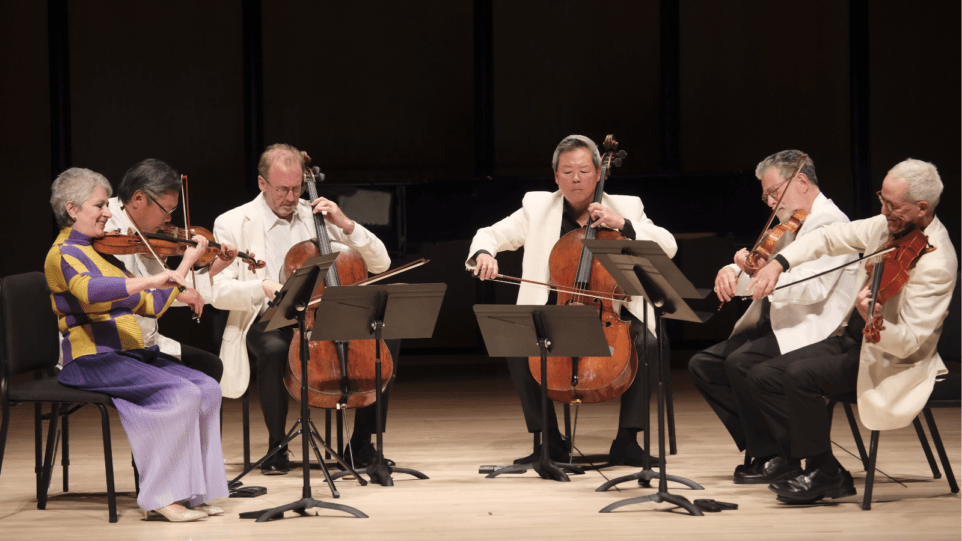 A group of six string players sitting in a semi-circle, performing a string sextet by Brahms. The five men are in white suit jackets. The one woman is the first chair violinist and dressed in a purple skirt with a yellow and purple sweater top.