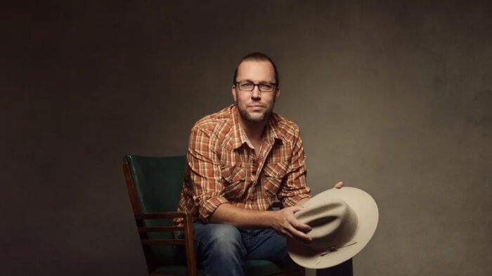 seated man wearing plaid shirt and holding his hat looks into the camera