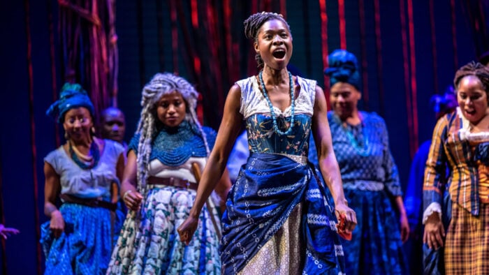 A woman in customary dress sings onstage with cast in the background