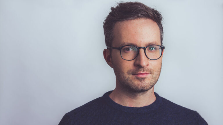 Half-smiling Conor Hanick in glasses and a blue sweater in front of a light blue background