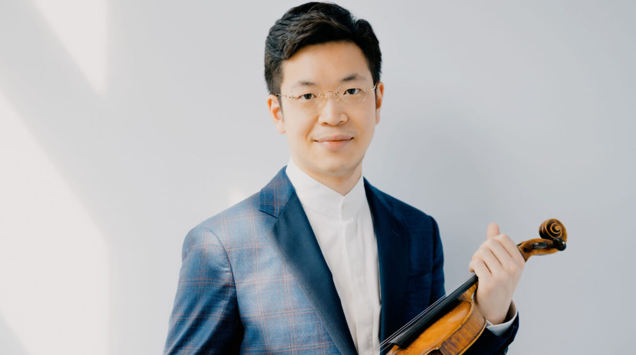 Paul Huang stands against a sunlit wall, wearing a blue sportcoat, holding a violin
